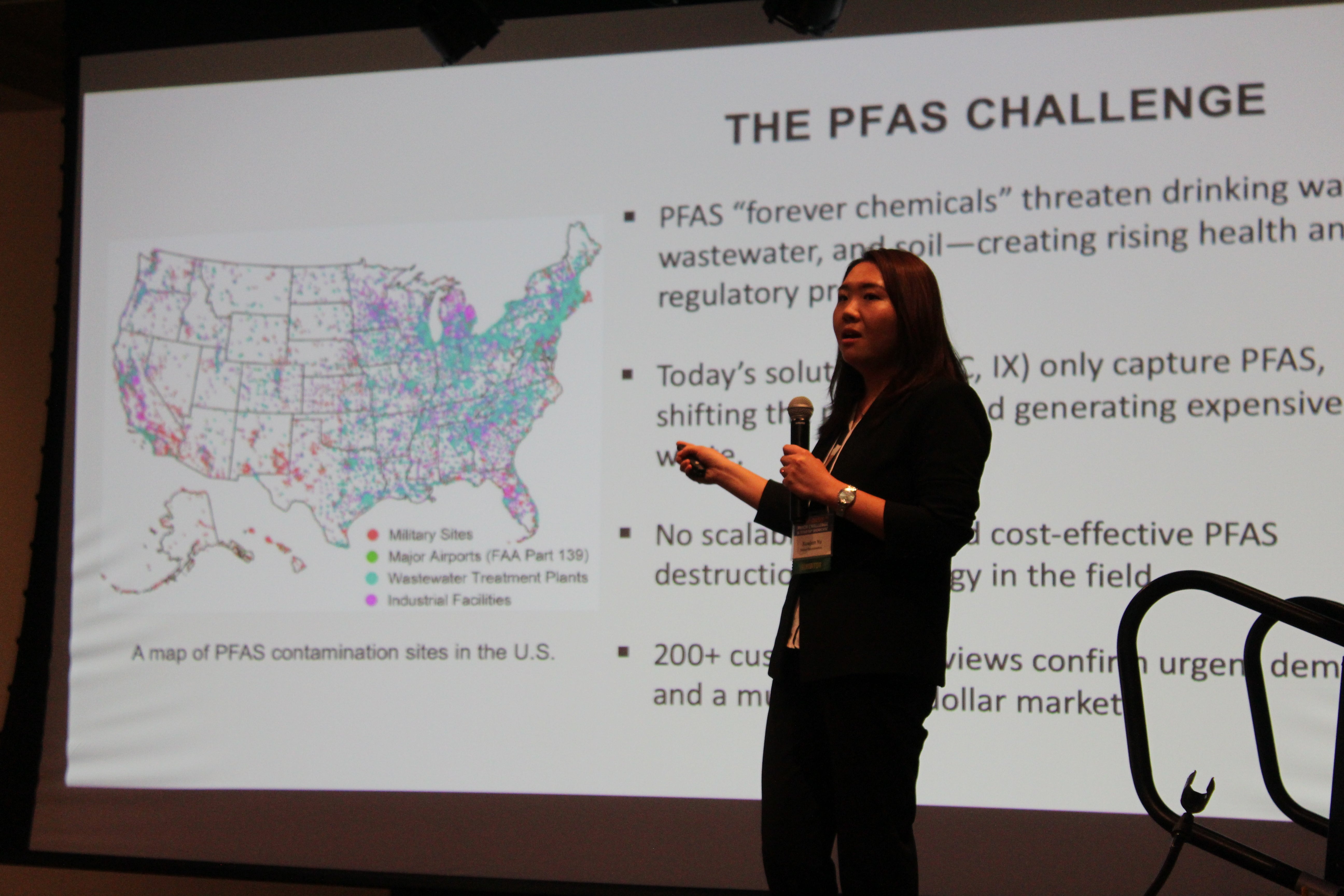 A woman on stage presenting her business pitch for her startup company in front of a large screen.